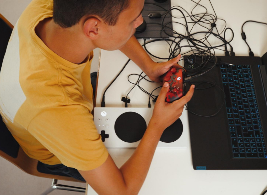 Jugendlicher zockt mit herkömmlichen XBox-Controller, vor ihm ein XBox-Adaptive-Controller.