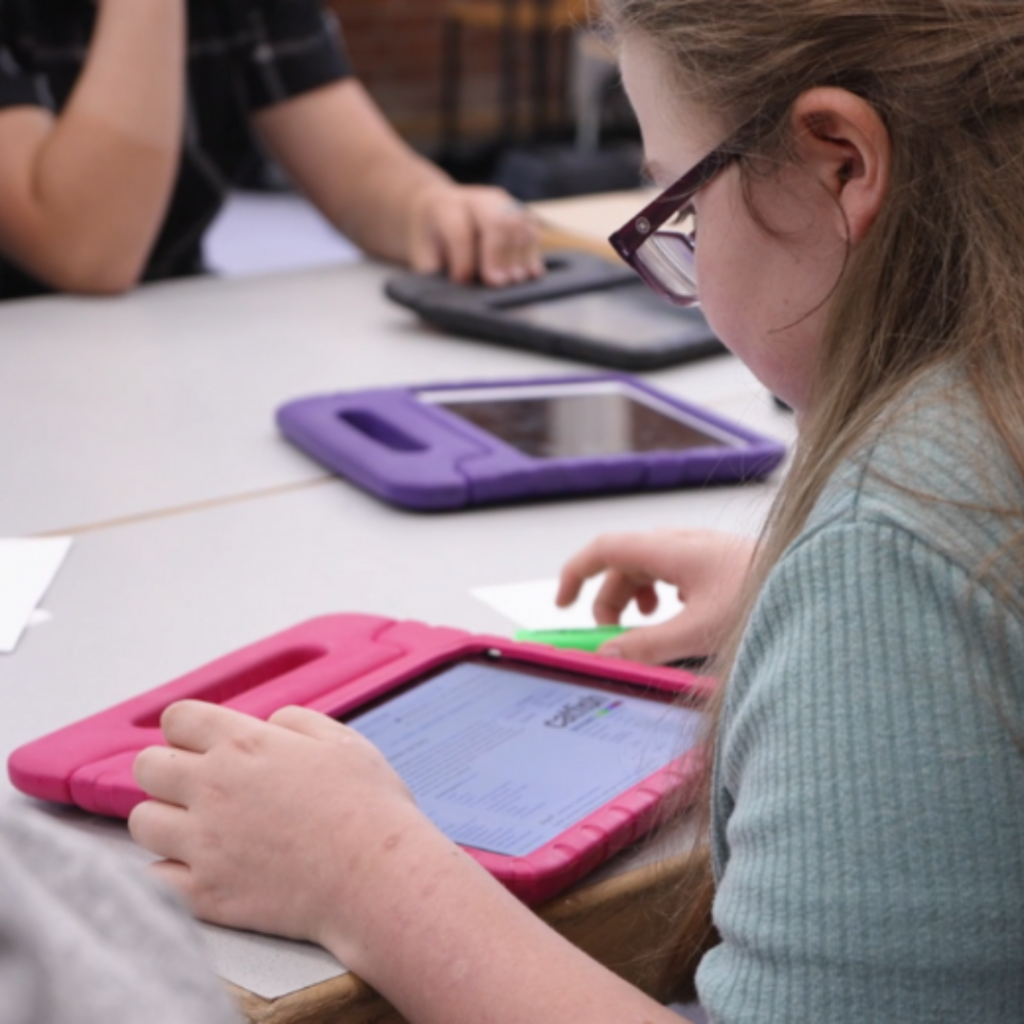 Jugendliche sitzt an einem Tisch. Auf dem Tisch liegen verschiedene Tablets, Stifte und Papier